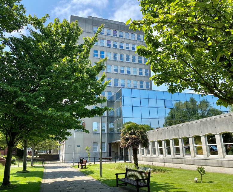 An image of Folkestone Civic Centre in late Spring surrounded by green grass and full trees as well as a row of benches